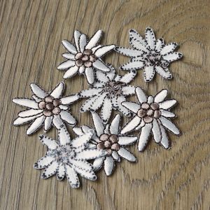 Mehrere Edelweiss Stick-Applikationen zum Aufbügeln (Ø 3 cm) in Offwhite/Braun, locker arrangiert auf einem Holz-Hintergrund. Vorder- und Rückseitenansichten.