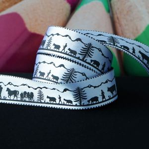 Nahaufnahme des glänzenden Webbands "Scherenschnitt Alpaufzug" (16 mm breit, silberweiss-schwarz) auf einem Stoffstück, die detaillierte Musterung ist gut sichtbar.